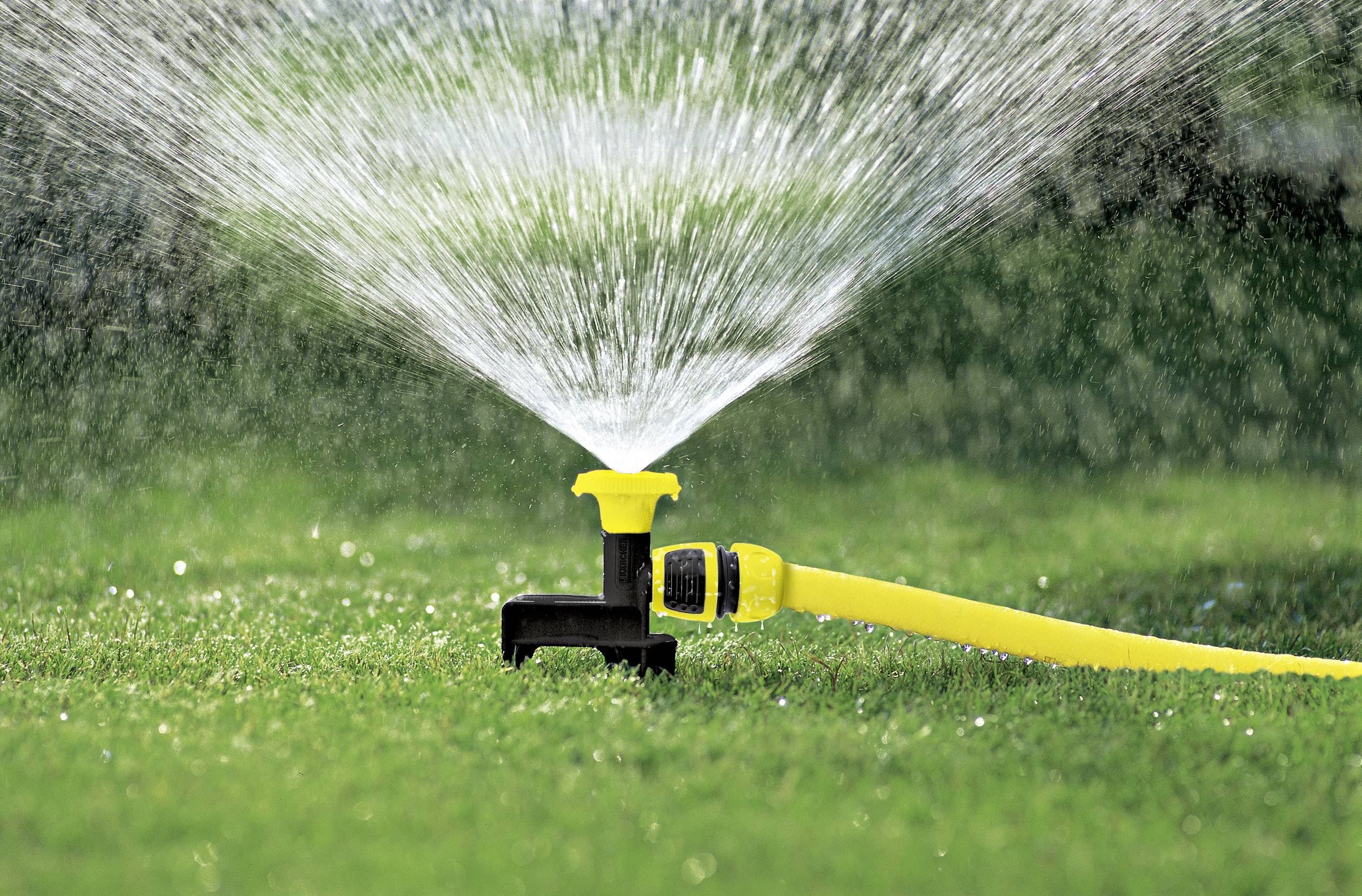 Un arroseur jaune répand uniformément de l'eau sur un gazon vert qui brille sous la lumière du soleil.