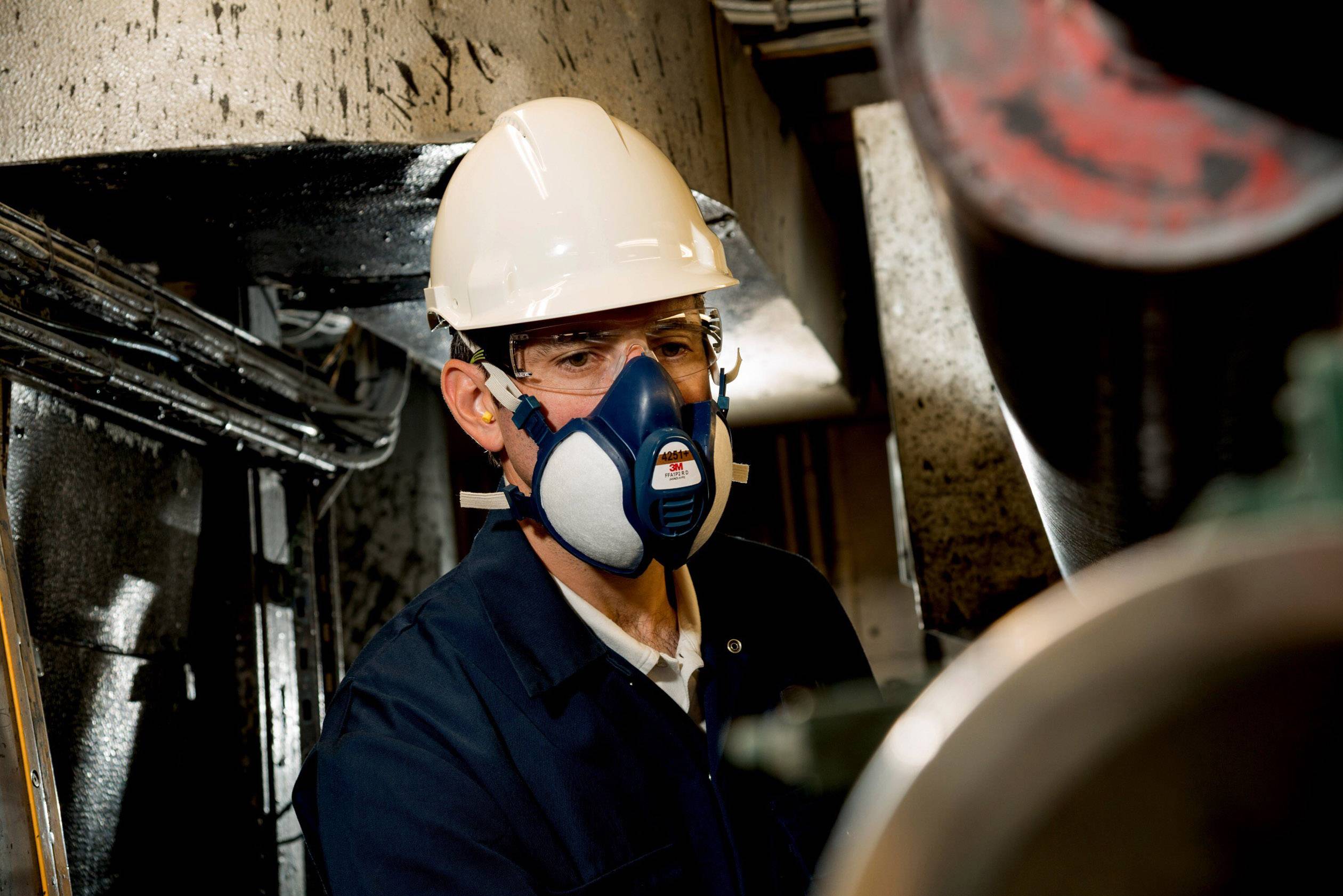 Une personne portant un casque de protection et un masque travaille dans un environnement industriel. L'accent est mis sur la sécurité et les mesures de protection.