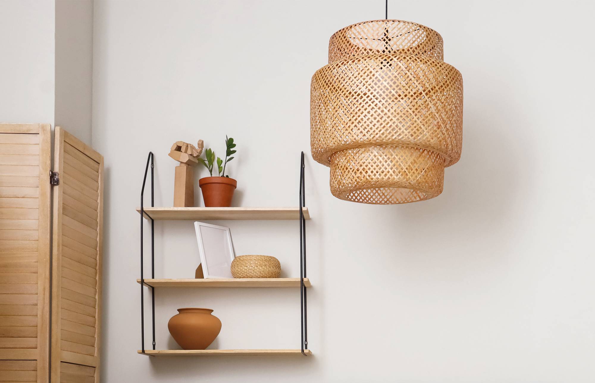 Coin de pièce minimaliste avec une suspension tissée au-dessus d'une étagère murale contenant une plante, un cadre, un panier et de la poterie, à côté d'un paravent en bois.