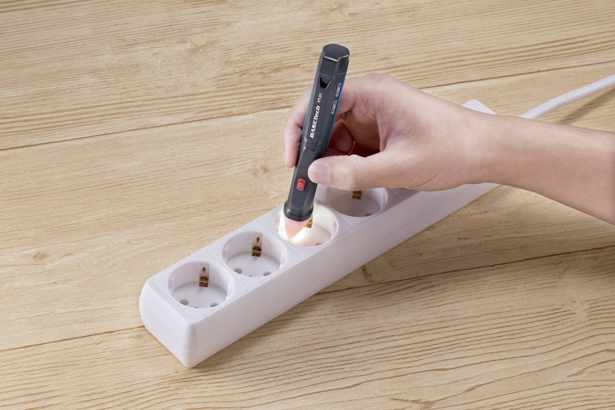 Une main tient un testeur de tension au-dessus d'une prise électrique dans une multiprise posée sur une table en bois, afin de vérifier la tension.