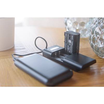 Un chargeur contenant deux batteries est connecté à une batterie externe via un câble USB sur une table en bois.
