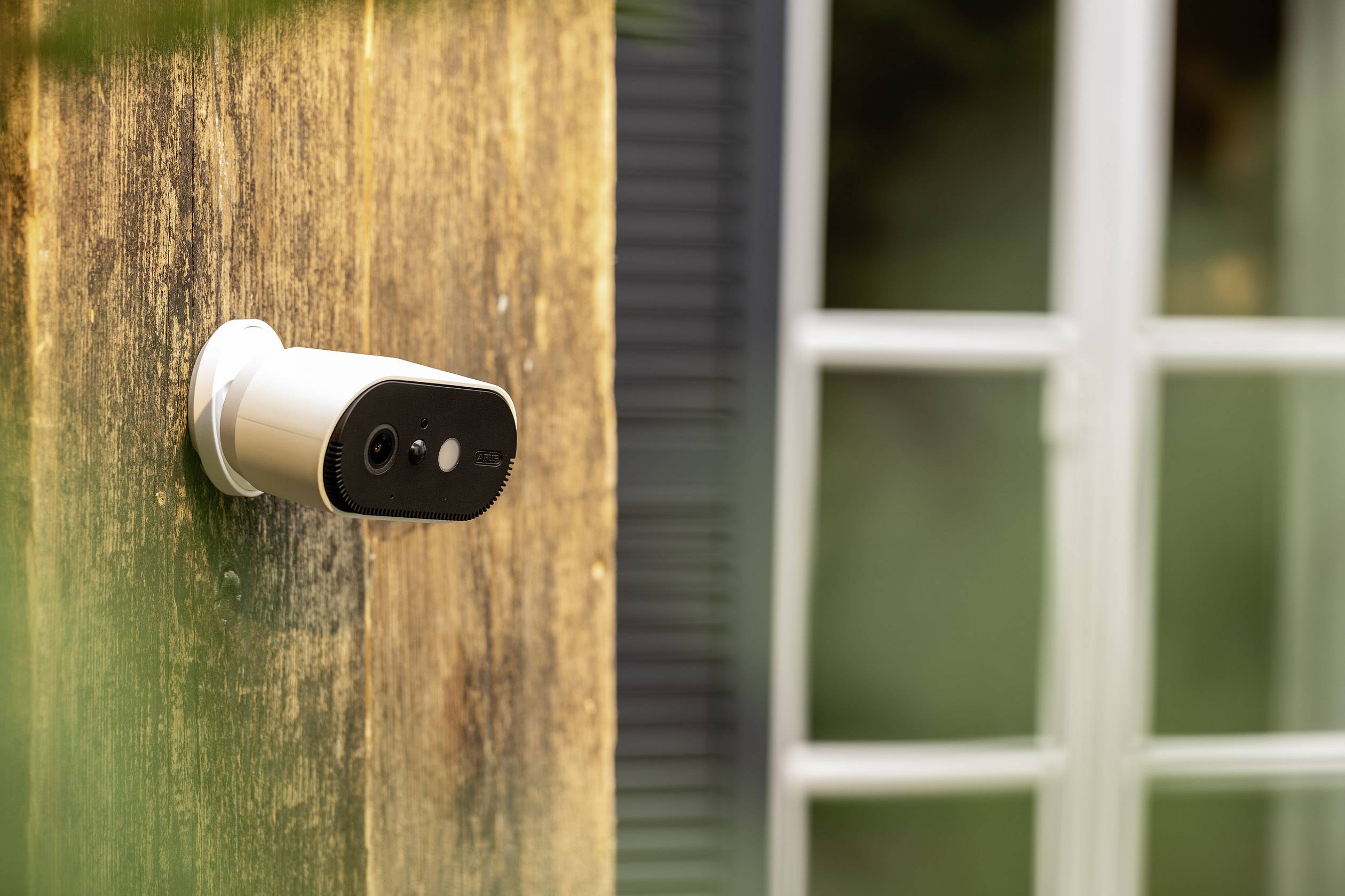 Une caméra de surveillance est installée sur le mur extérieur d'une maison, avec une fenêtre visible en arrière-plan.