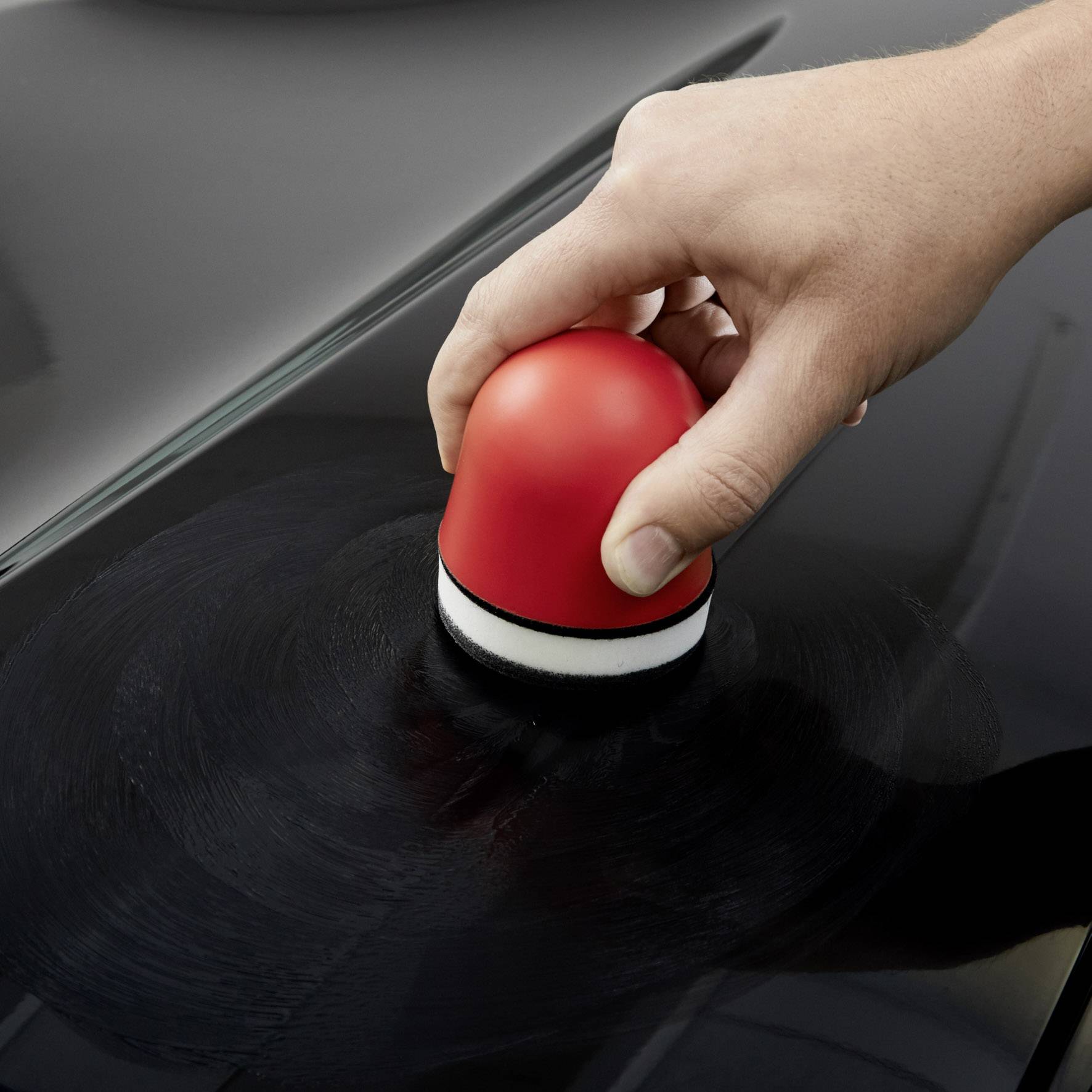 Une main polit une surface de peinture automobile noire avec un pad de polissage rouge. La surface effectue des mouvements circulaires.