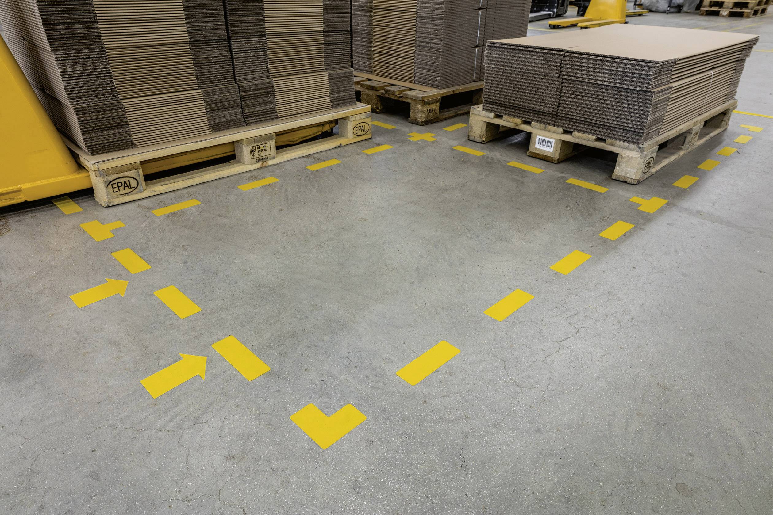 Des marquages jaunes sur le sol d'un entrepôt indiquent une zone de circulation sécurisée. Des palettes chargées de cartons sont adjacentes.