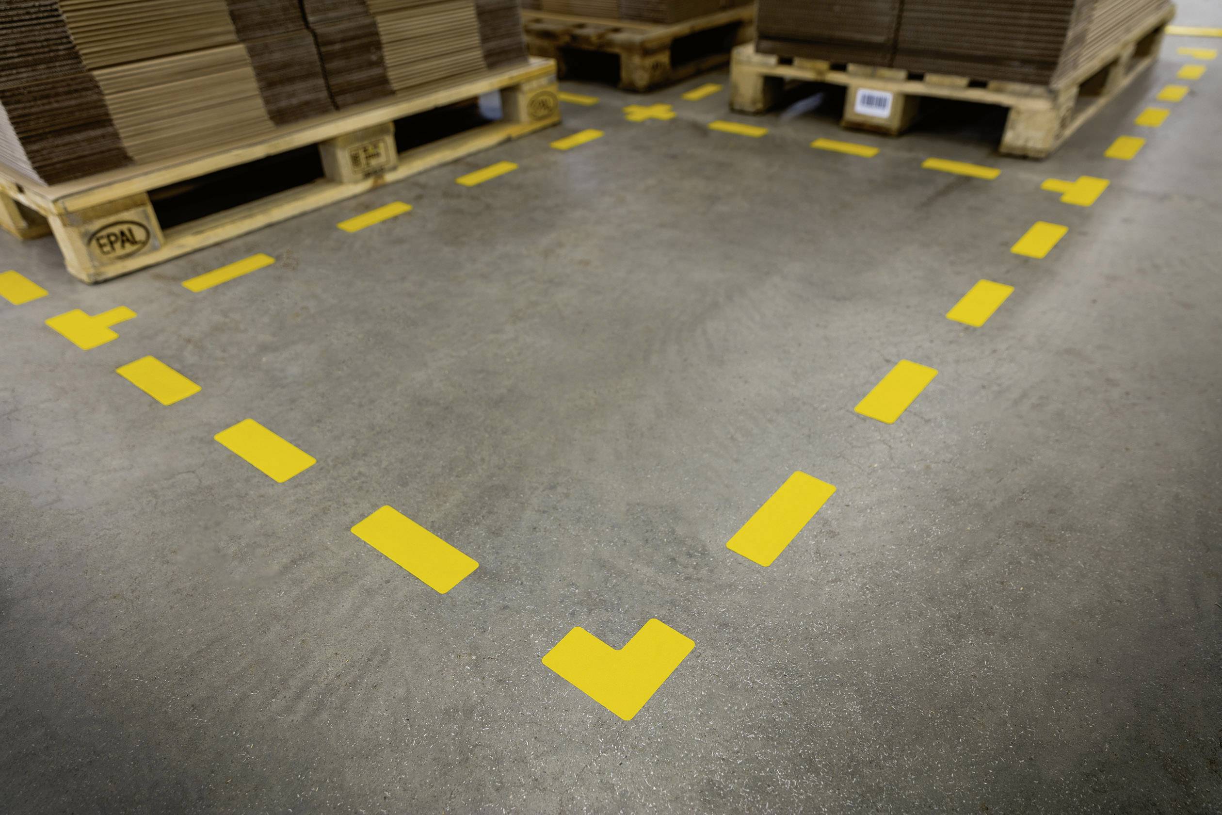 Des palettes chargées de cartons sont empilées dans un entrepôt. Des marquages jaunes au sol délimitent la zone.