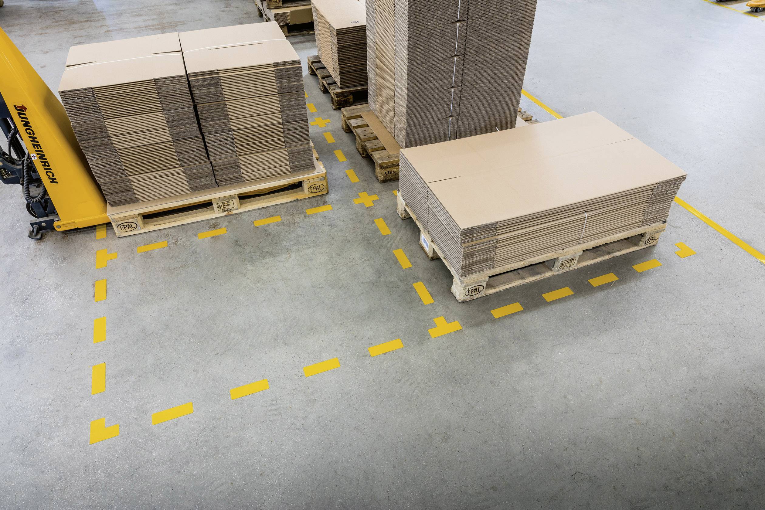 Plusieurs cartons empilés sur des palettes dans une zone d'entreposage, délimités par des lignes jaunes au sol pour l'organisation et la sécurité.