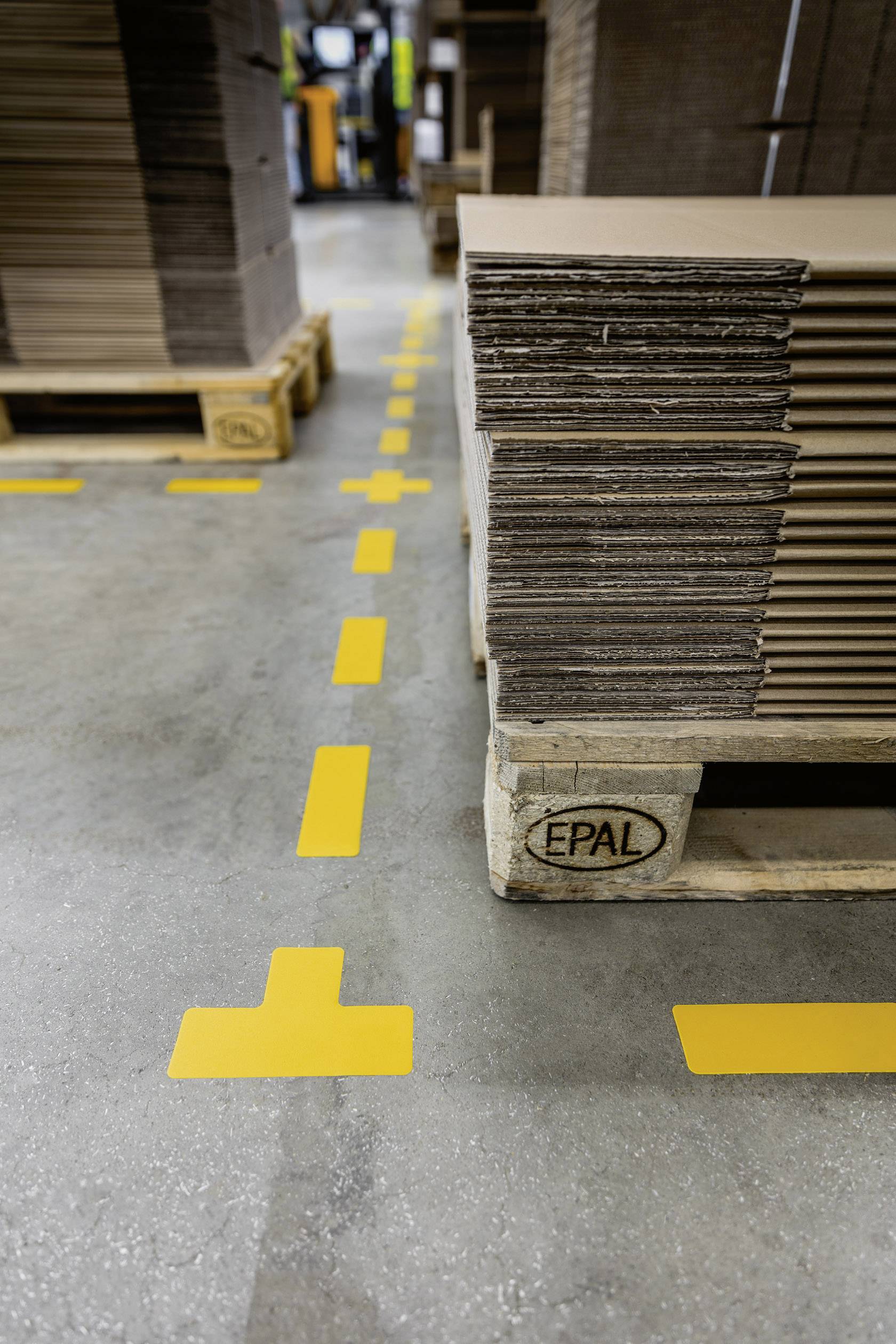 Palette de cartons empilés dans un entrepôt. Des marquages jaunes au sol indiquent les voies de circulation.