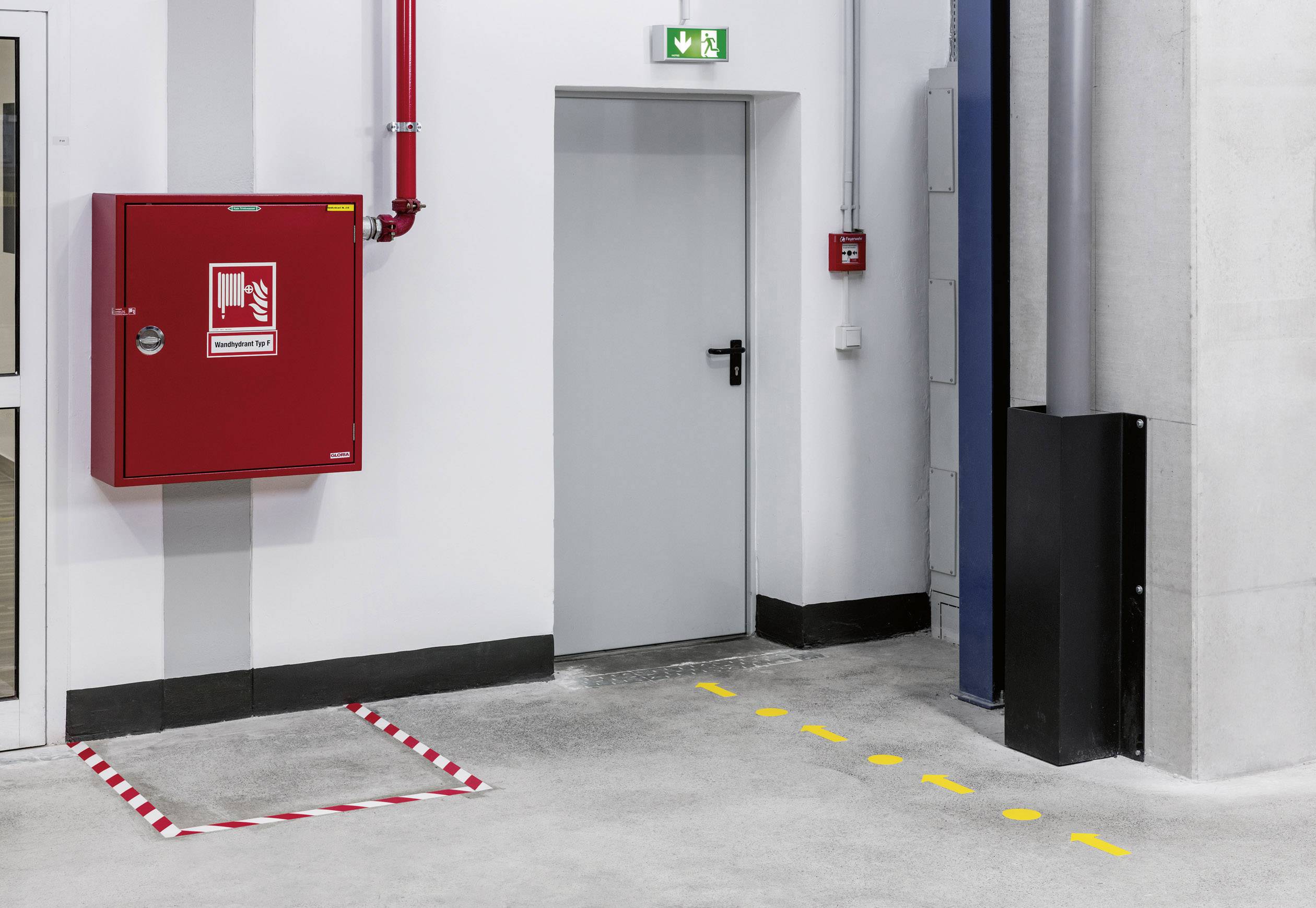 Une porte de sortie de secours dans un entrepôt avec un panneau de signalisation de sortie de secours vert au-dessus. À côté, un boîtier d'extincteur rouge fixé au mur.