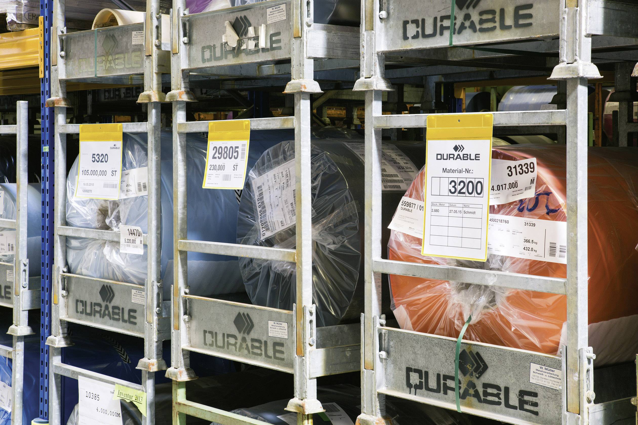 Différents rouleaux de matériaux colorés sont rangés sur une étagère. Des étiquettes indiquent les codes et numéros de matériaux pour l'organisation.
