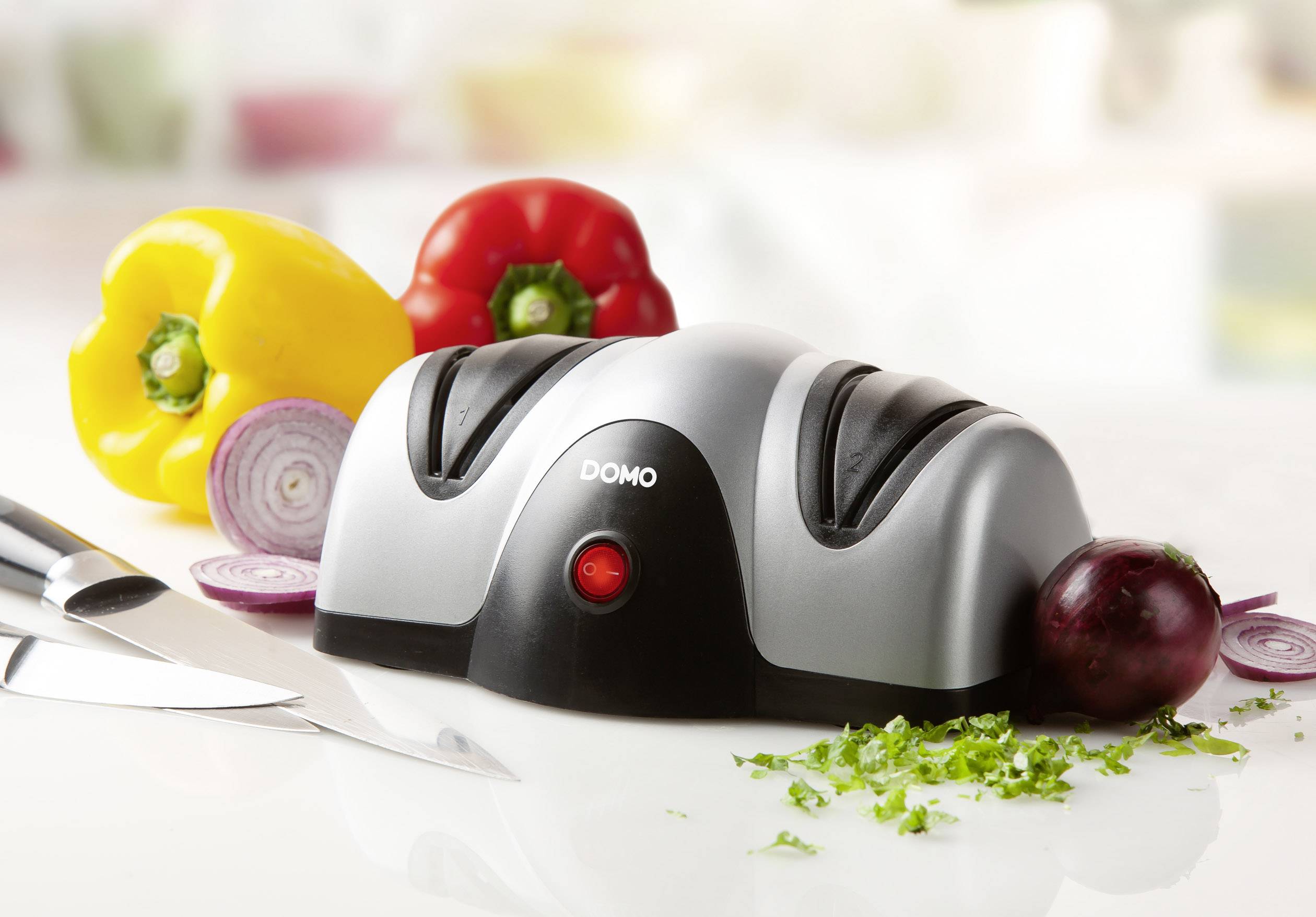 Aiguiseur de couteaux électrique avec voyant rouge sur une table de cuisine, entouré de poivrons, oignons et un couteau.