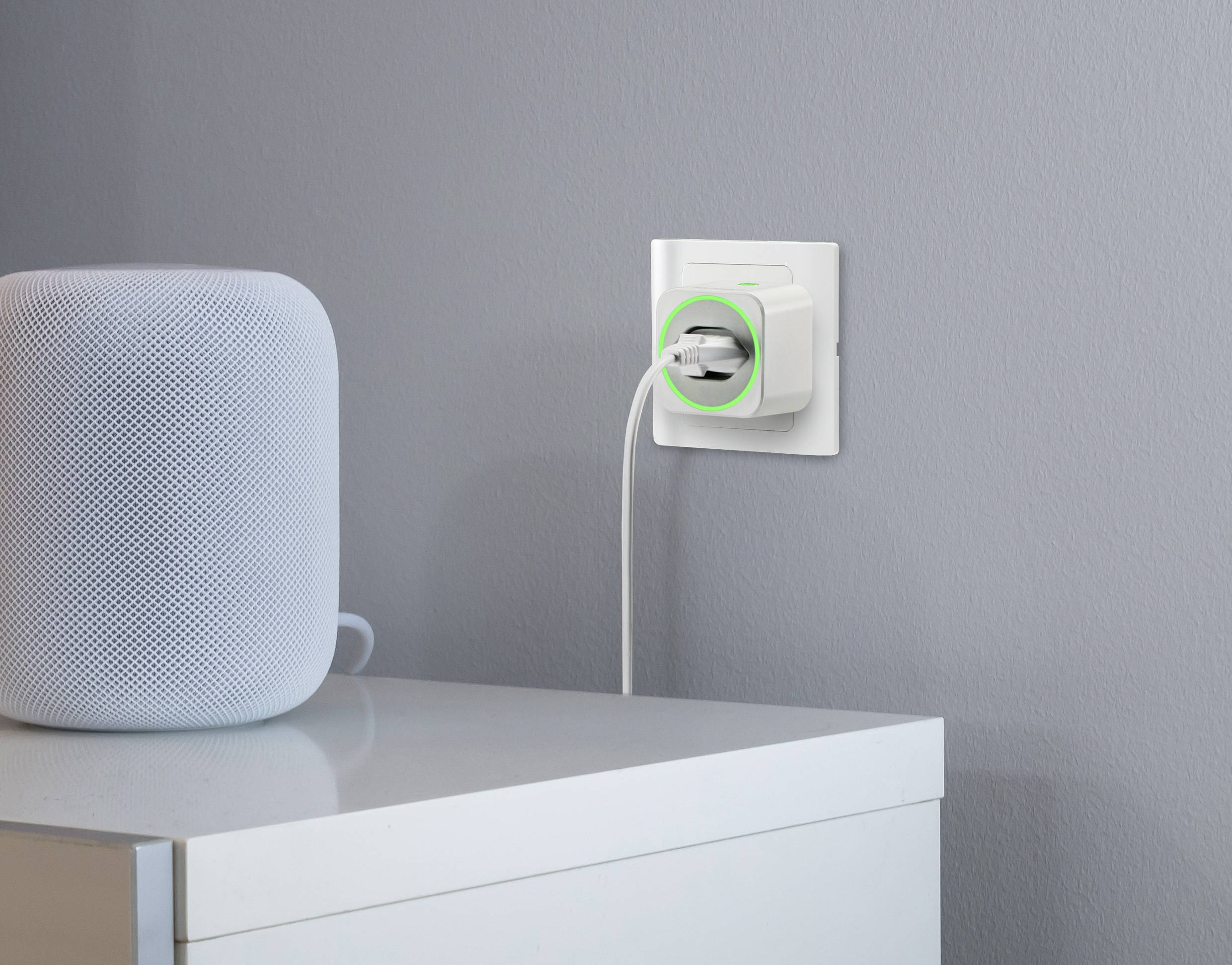 Prise électrique blanche sur un mur gris, un appareil domotique avec un anneau lumineux vert. À côté, sur une table blanche, se trouve un haut-parleur.