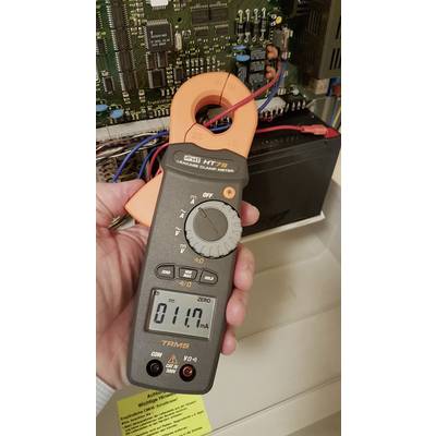 Une personne tient un ampèremètre numérique à pince qui affiche 0,117 ampère. En arrière-plan, un circuit électrique ouvert est visible.