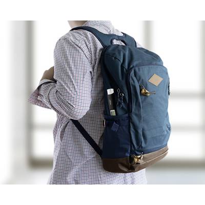 Une personne porte un sac à dos bleu. Le sac à dos possède plusieurs poches et des accents marron. Des vitres sont visibles en arrière-plan.
