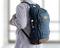 Une personne porte un sac à dos bleu. Le sac à dos possède plusieurs poches et des accents marron. Des vitres sont visibles en arrière-plan.