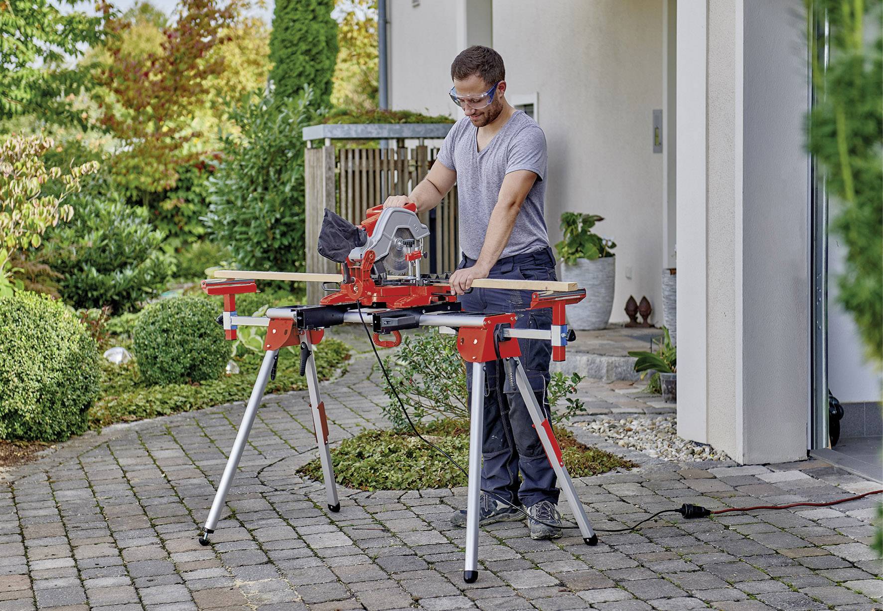 Un homme utilise une scie circulaire sur une table à l'extérieur sur une terrasse pavée pour couper une planche en bois.