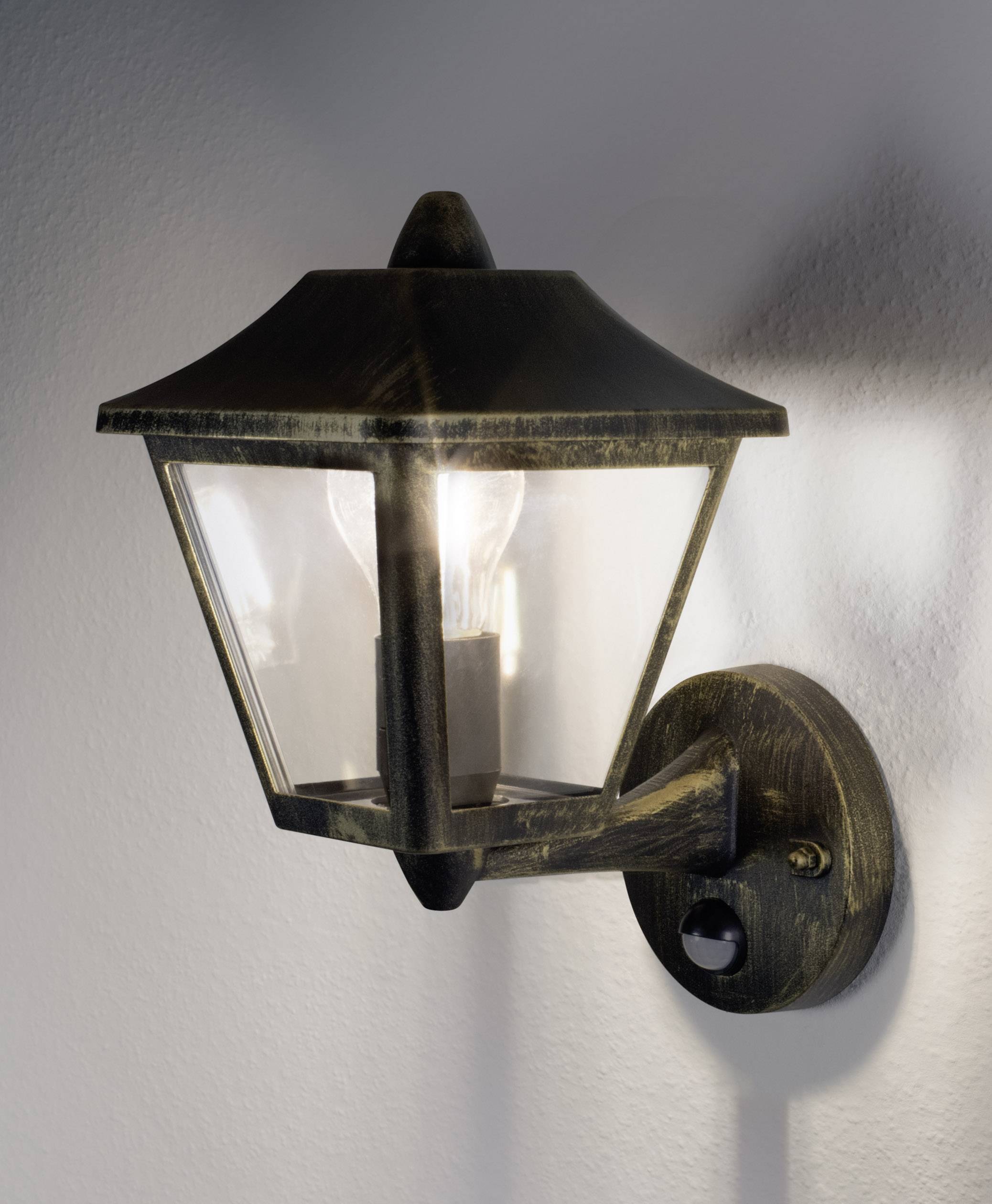 Applique murale au design antique avec un verre transparent, installée sur un mur. La lampe est allumée et diffuse une lumière chaleureuse.