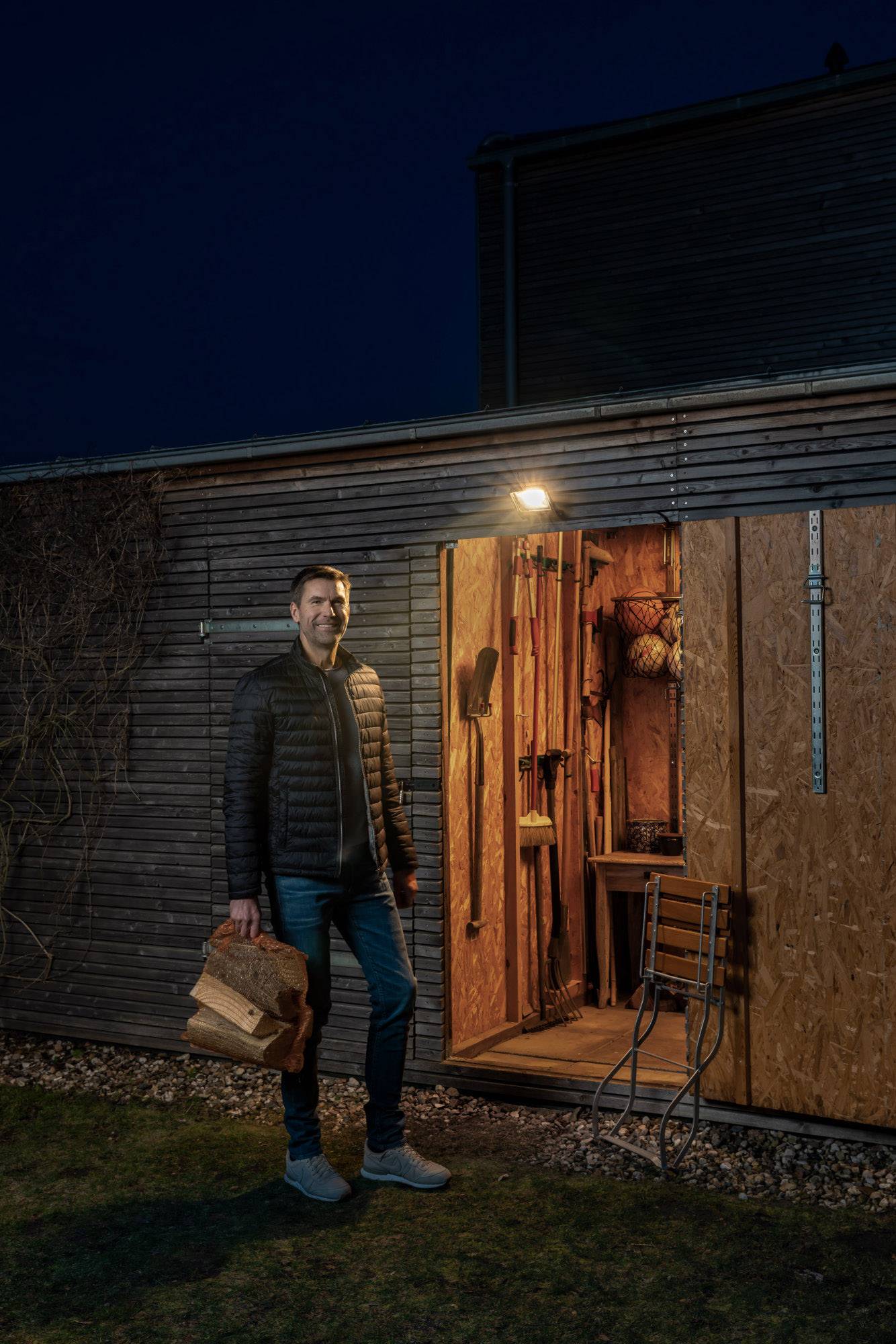 Un homme se tient devant une cabane en bois ouverte dans la nuit, tenant des bûches dans sa main gauche. La cabane est éclairée à l'intérieur.