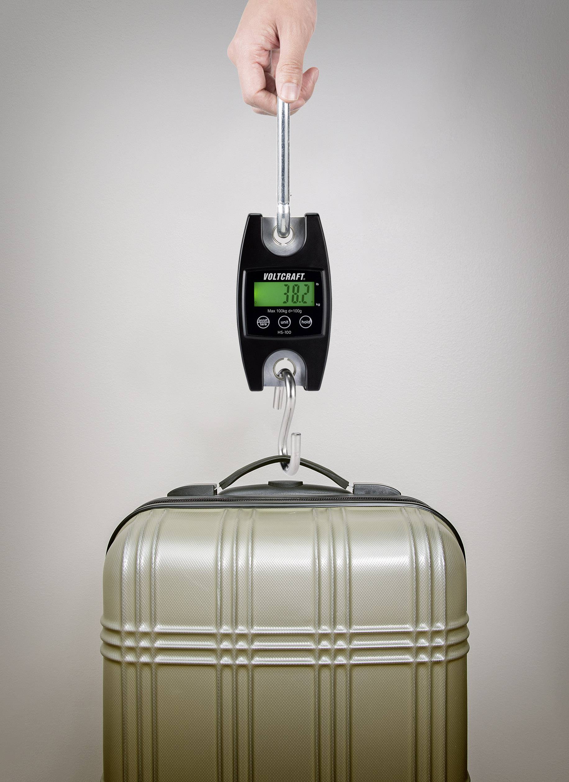 Une main tient une balance à bagages affichant 38,2 kg. La valise en dessous est de couleur argent et au design moderne.