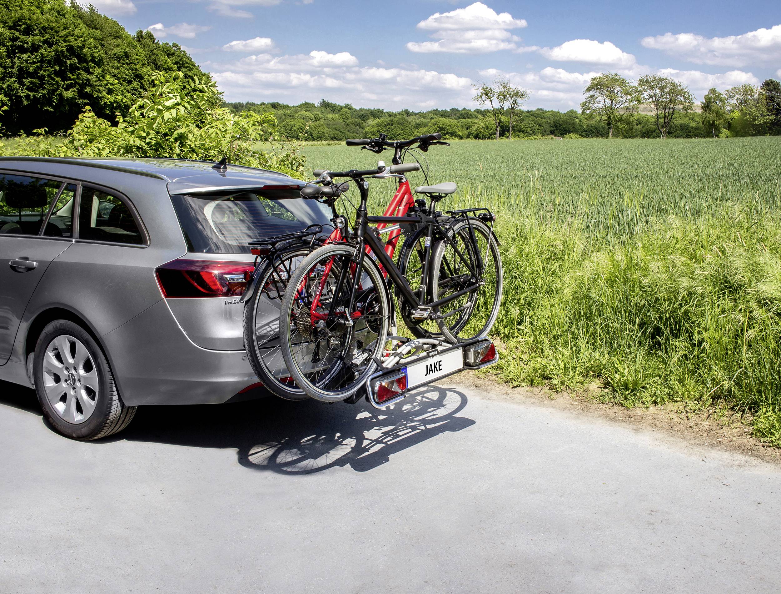 Voiture avec deux vélos sur un porte-vélos arrière, garée à côté d'un champ vert sous un ciel bleu avec quelques nuages.