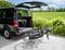 Un fourgon noir ouvert, stationné sur un chemin de campagne, montre deux vélos fixés à l'arrière, avec un champ vert en arrière-plan.