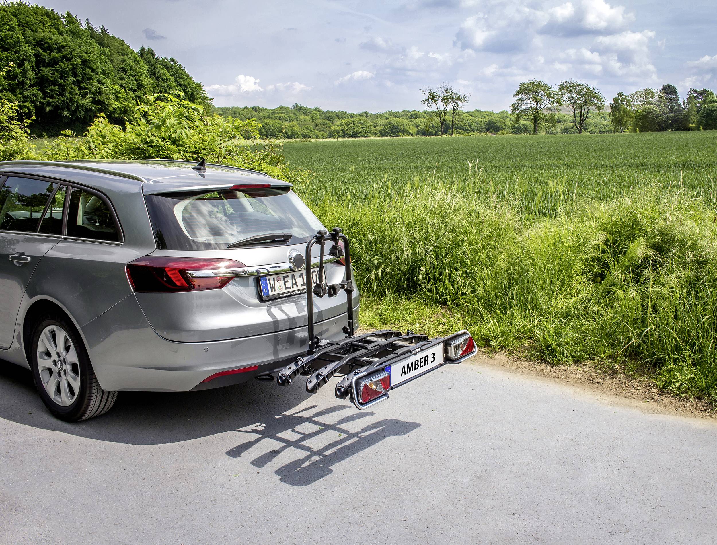 Une voiture argentée est garée sur un chemin de campagne, équipée d'un porte-vélos à l'arrière, entourée de champs verts et d'arbres en arrière-plan.