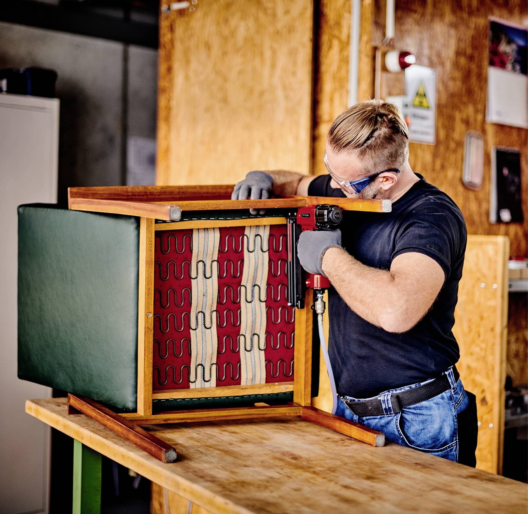 Une personne répare une chaise alors qu'elle est posée sur un établi. Elle porte des lunettes de protection et des gants dans un environnement d'atelier.