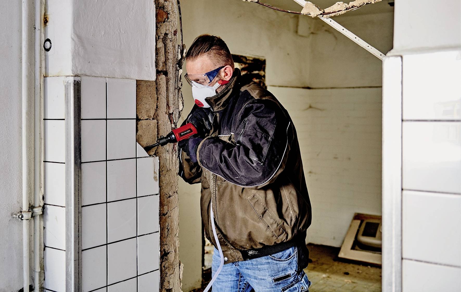 Une personne portant un masque de protection retire des carreaux d'un mur dans un bâtiment, probablement dans le cadre de travaux de rénovation.