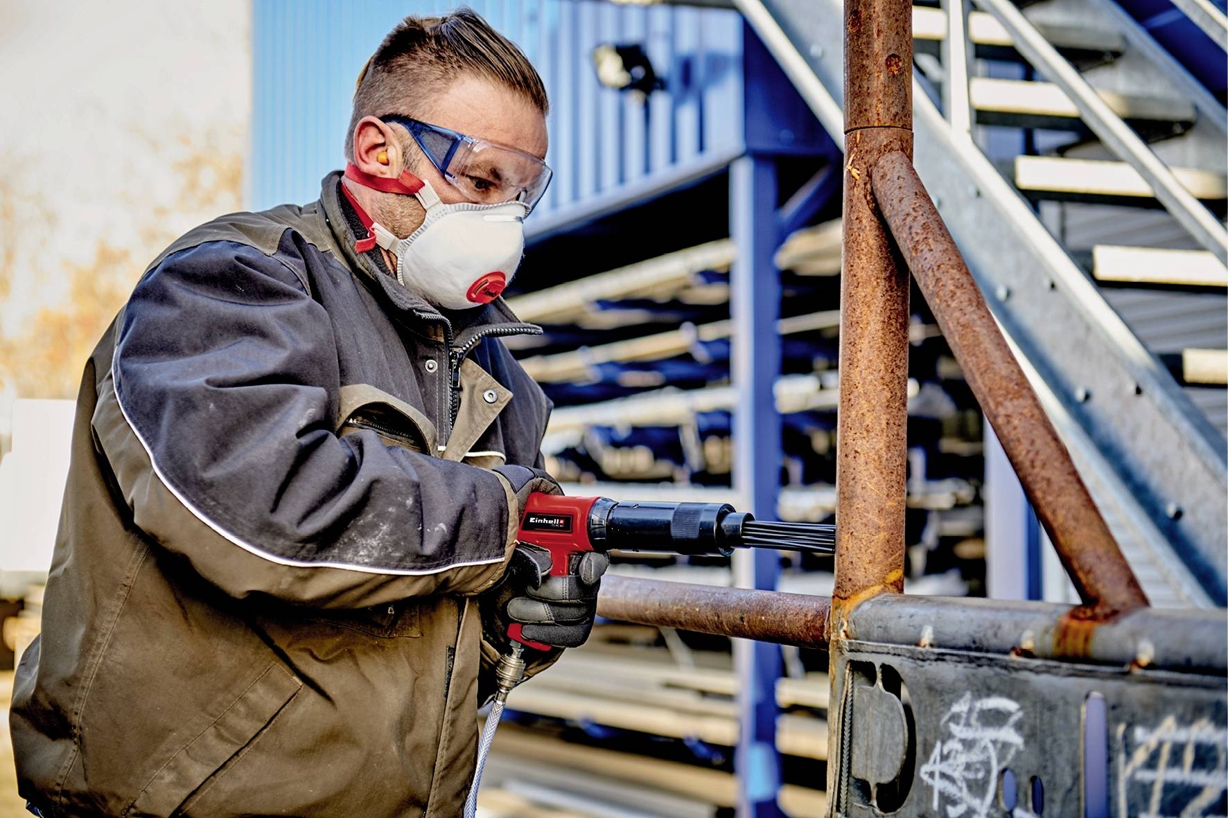 Un ouvrier portant des lunettes de protection et un masque respiratoire utilise un outil électrique pour travailler une rambarde métallique rouillée à l'extérieur.