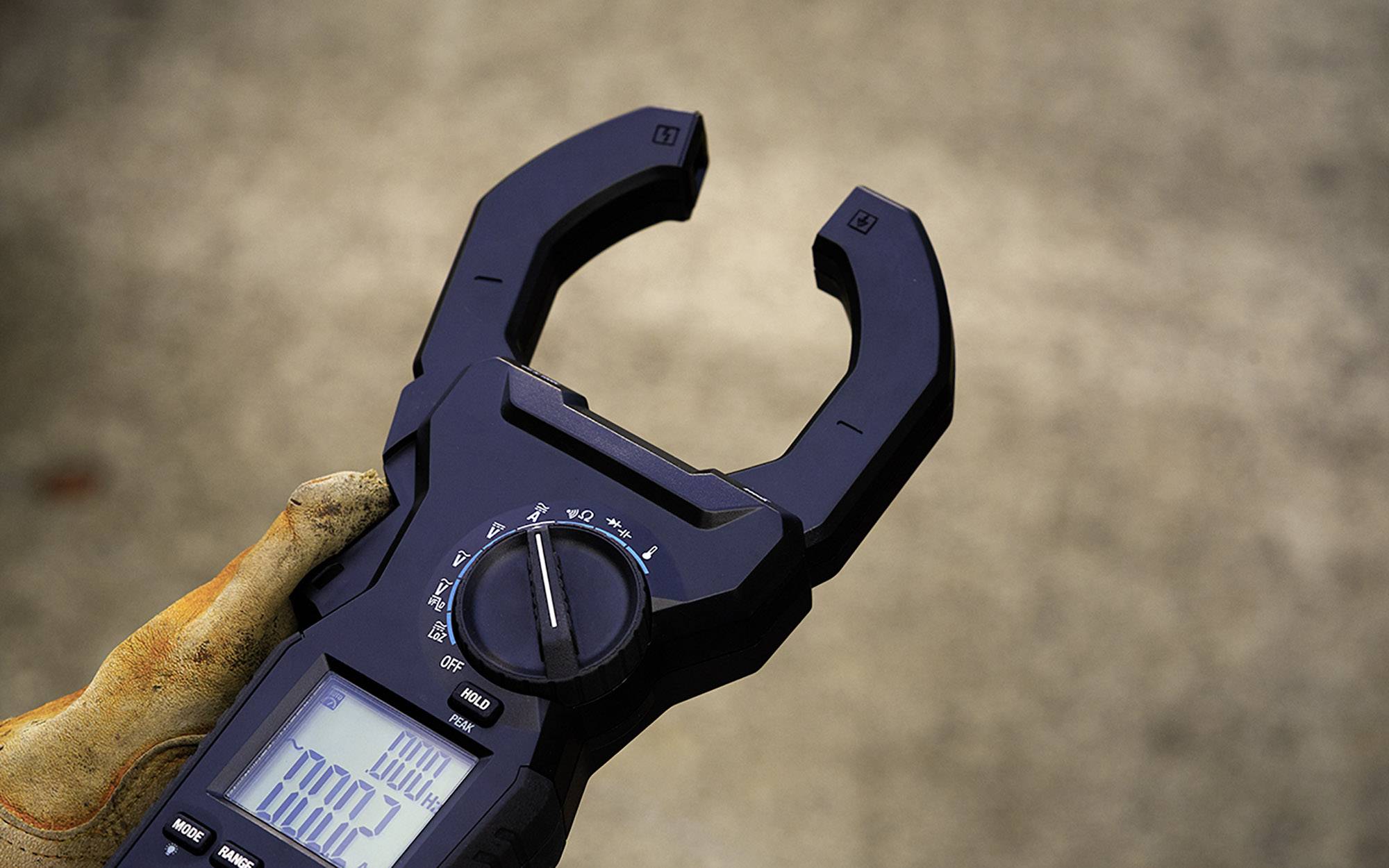 Un multimètre est tenu par une main gantée, affichant des valeurs de mesure numériques. Gros plan sur un fond gris.