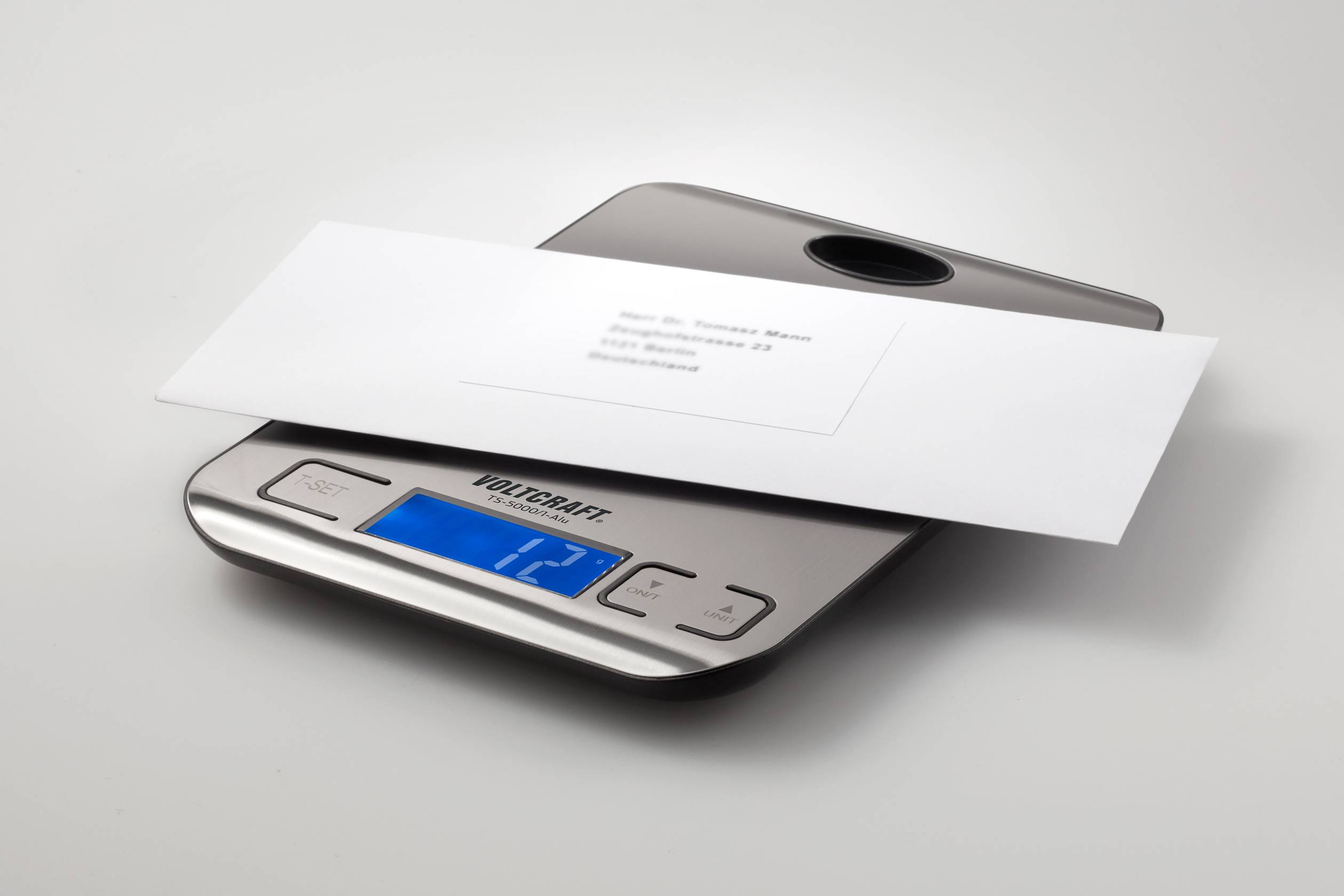 Une enveloppe blanche est posée sur une balance numérique. L'affichage indique un poids de 12 grammes.