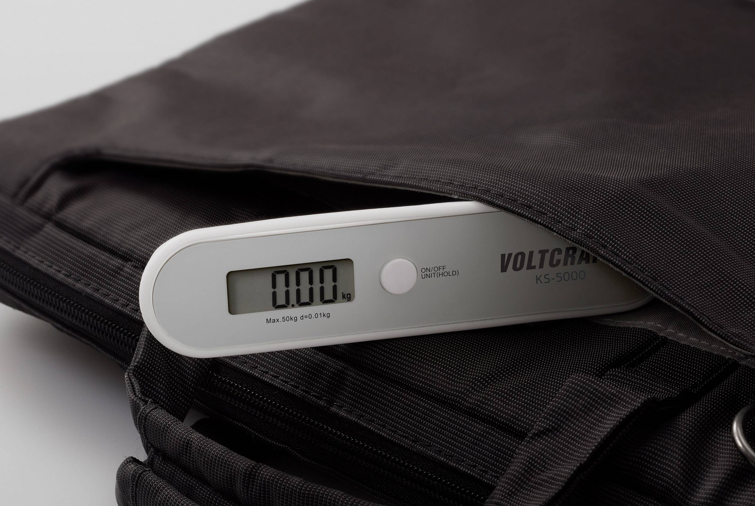 Balance numérique avec écran affichant 0,00 kg. L'appareil est posé sur un sac noir. Le nom de marque 'Voltcraft' est visible.