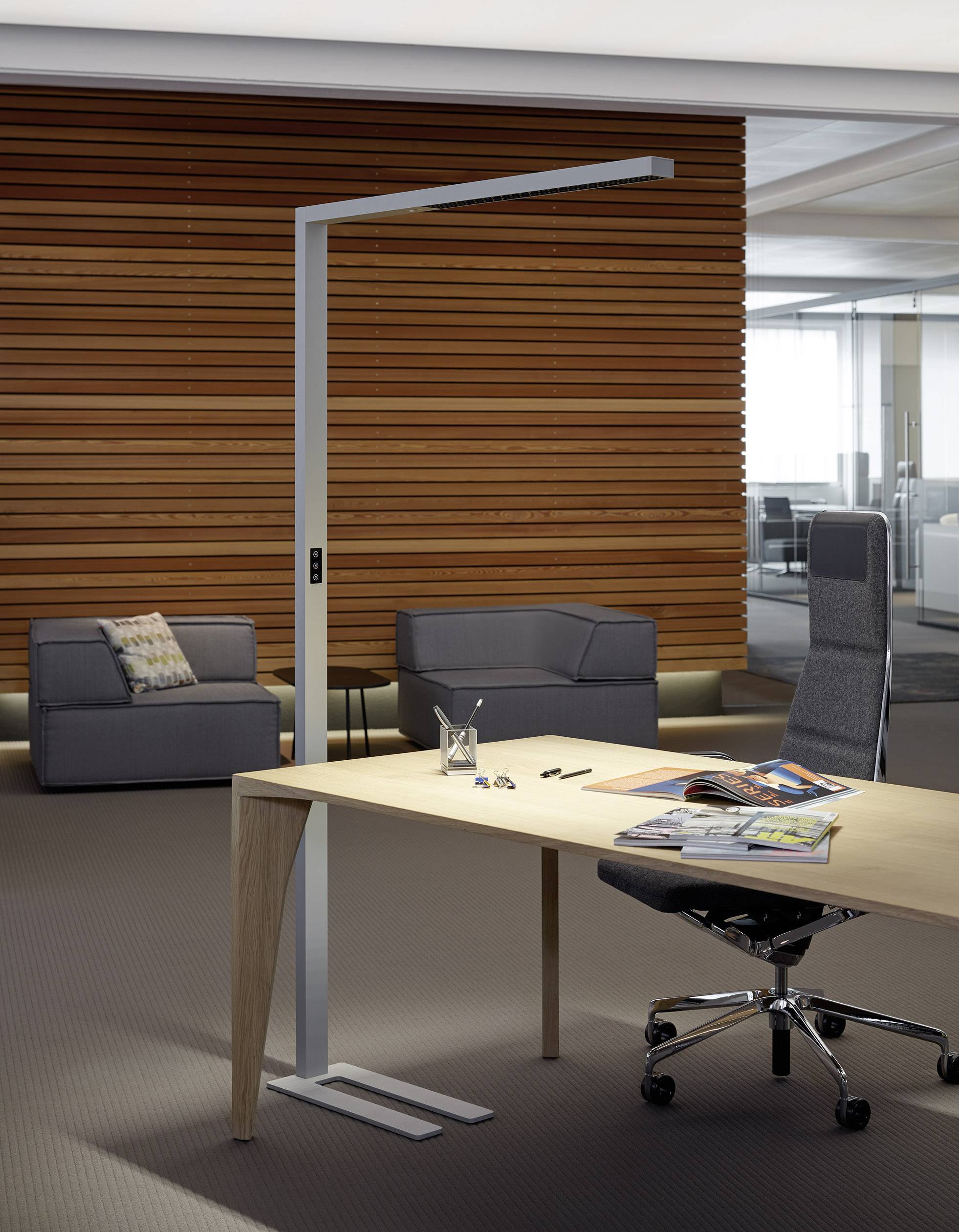 Espace de bureau moderne avec mur en bois : bureau avec une chaise de haute qualité et une lampe sur pied, deux fauteuils gris à l'arrière-plan, dossiers sur la table.