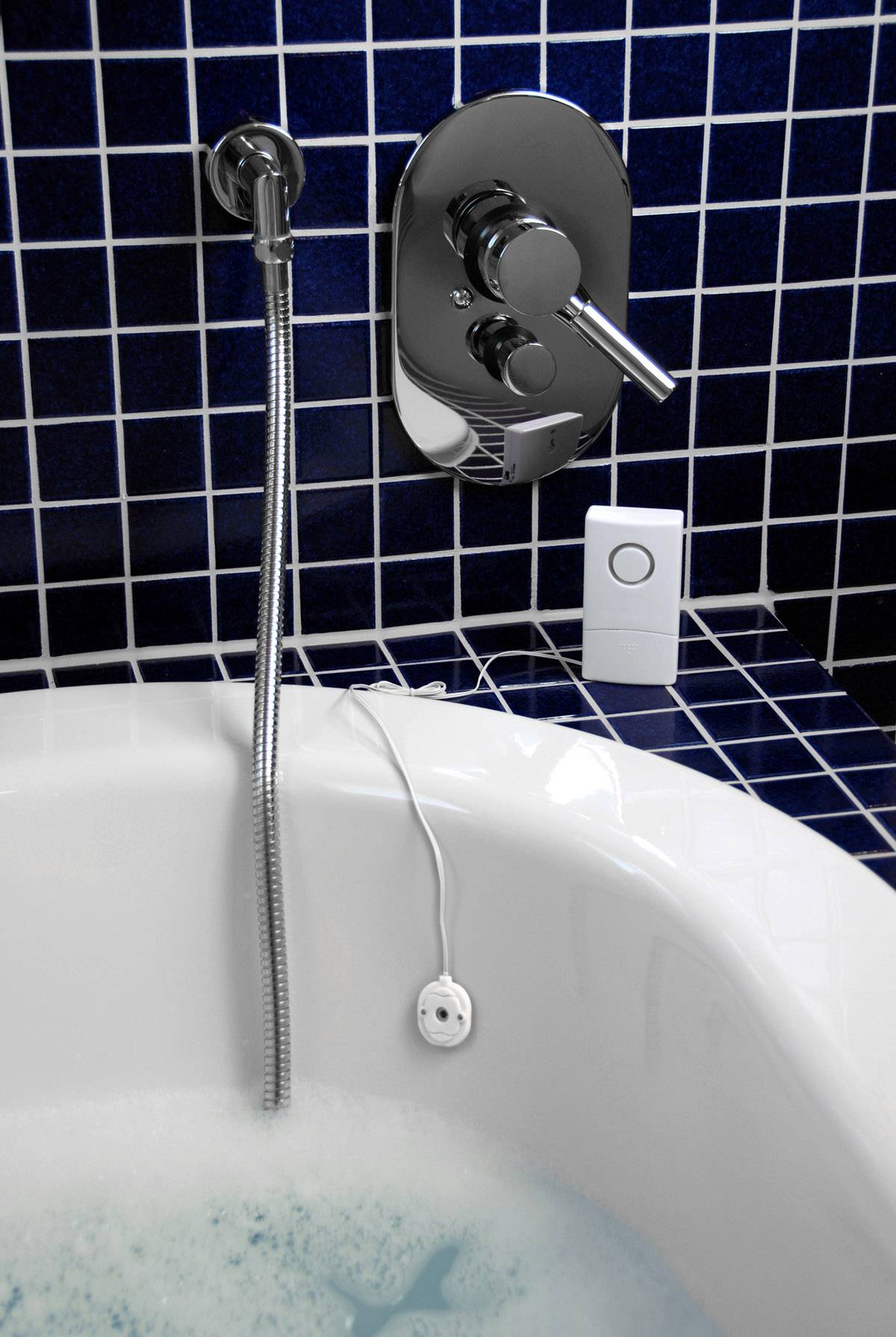 Baignoire remplie d'eau et de mousse, avec un capteur relié par un câble. Sur le mur, un pommeau de douche et un mitigeur chromé lisse, installés sur des carreaux bleus.