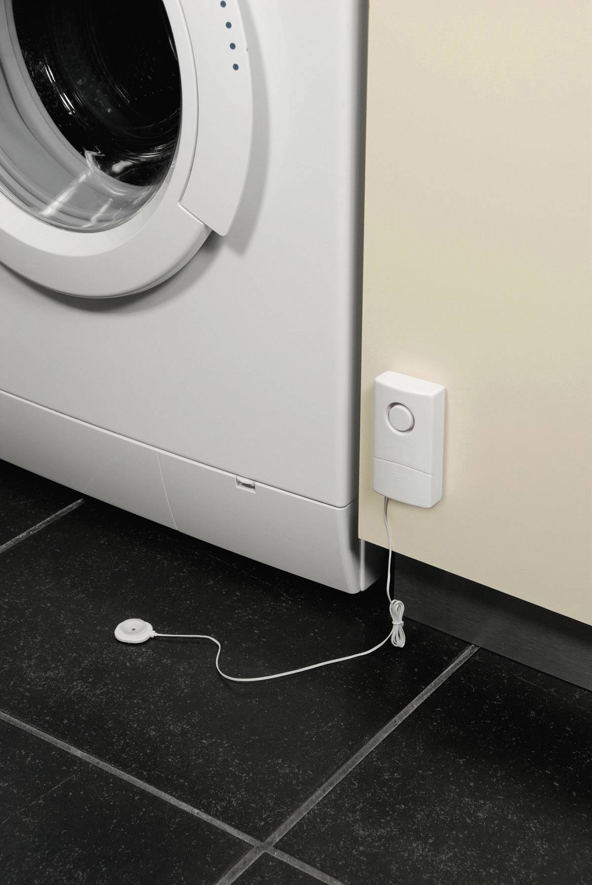 Une machine à laver à côté d'un détecteur d'inondation fixé au mur. Un capteur est placé sur le sol pour détecter les fuites.