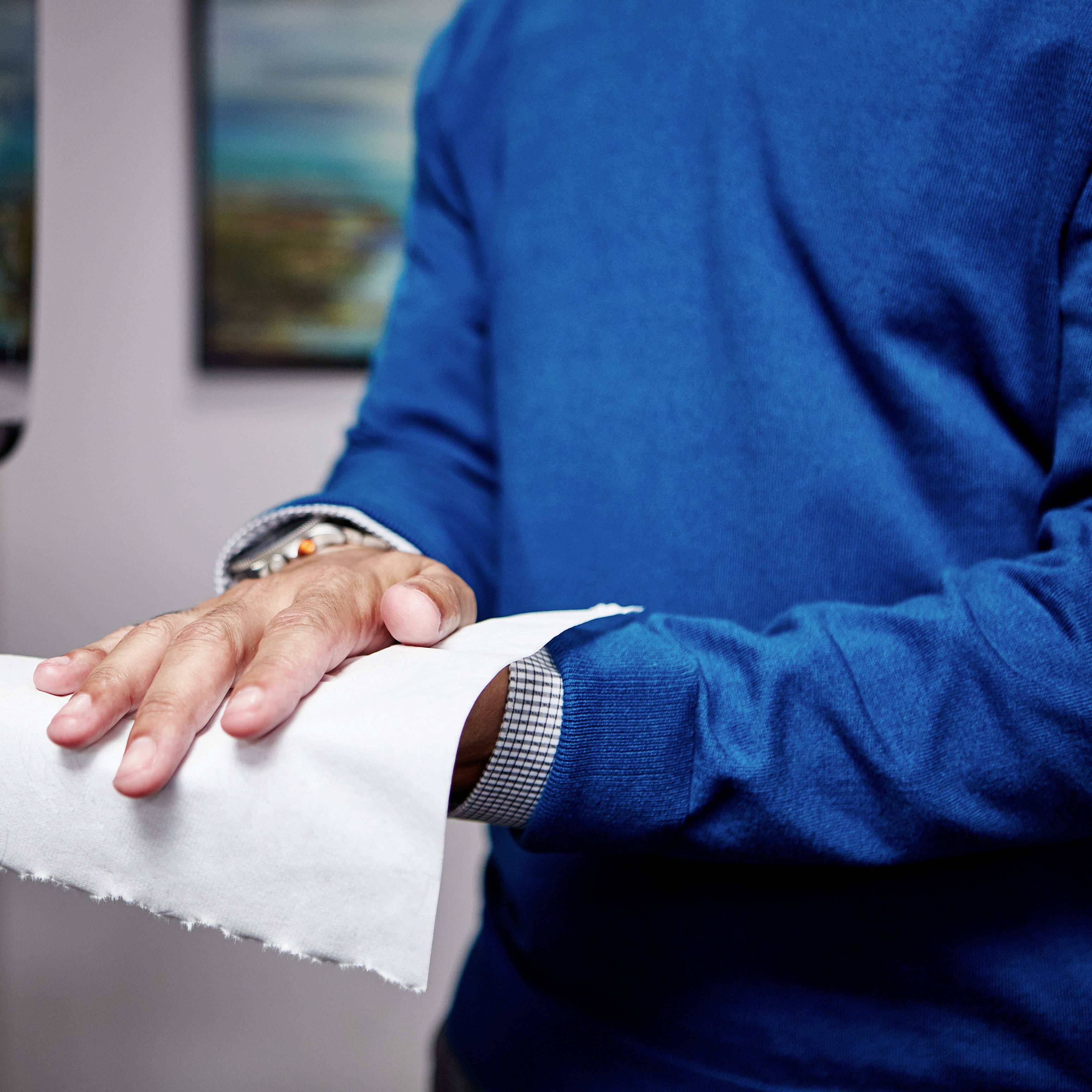 Une personne déchire une feuille de papier avec sa main droite au niveau de la bordure de couture. La personne porte un pull bleu.