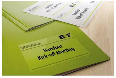 Pochette verte intitulée 'Handout Kick-off Meeting' posée sur une table, avec quelques feuilles volantes portant des notes.