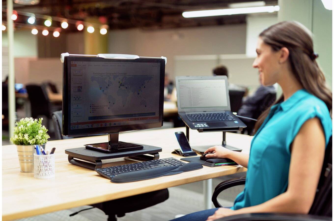 Une femme est assise dans un bureau moderne à un bureau équipé de deux écrans, d'un ordinateur portable et d'un smartphone. Elle travaille de manière concentrée sur son ordinateur.