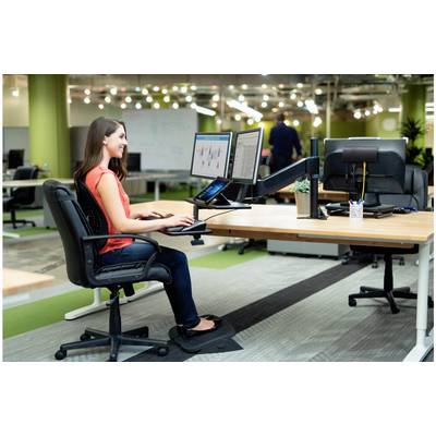 Une femme est assise à un bureau dans un bureau, utilisant deux écrans d'ordinateur. L'environnement est moderne et en open-space avec un éclairage lumineux.