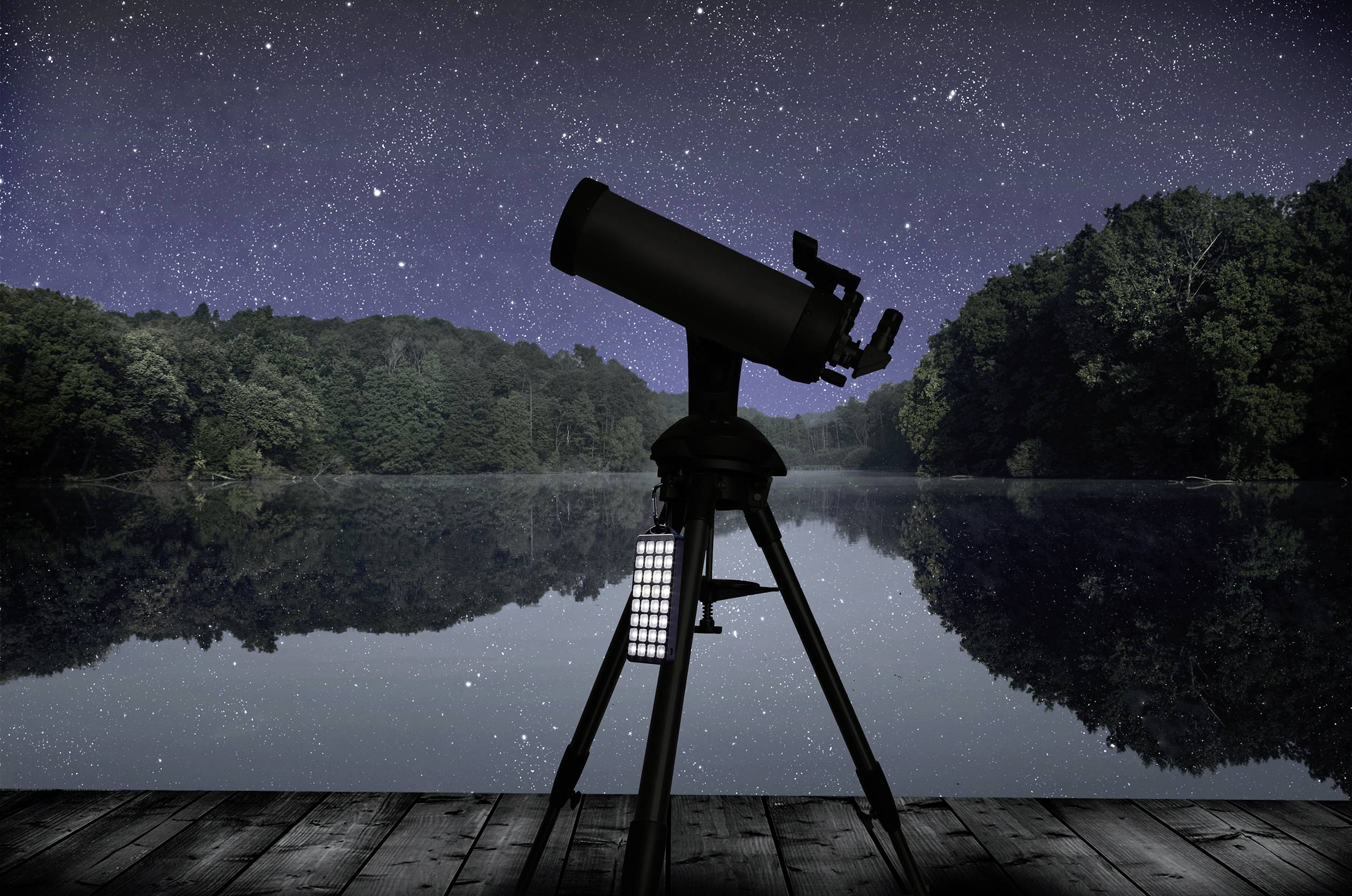 Un télescope est installé sur une terrasse en bois, pointé vers un ciel nocturne étoilé au-dessus d'un lac paisible bordé de forêts.