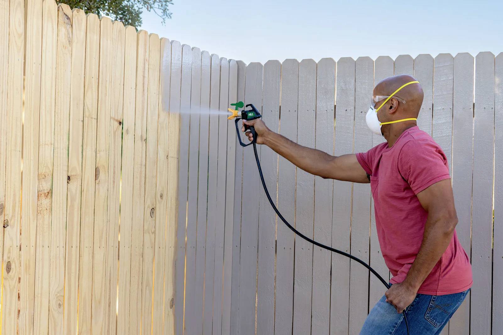 Un homme portant un masque de protection pulvérise de la peinture sur une clôture en bois.