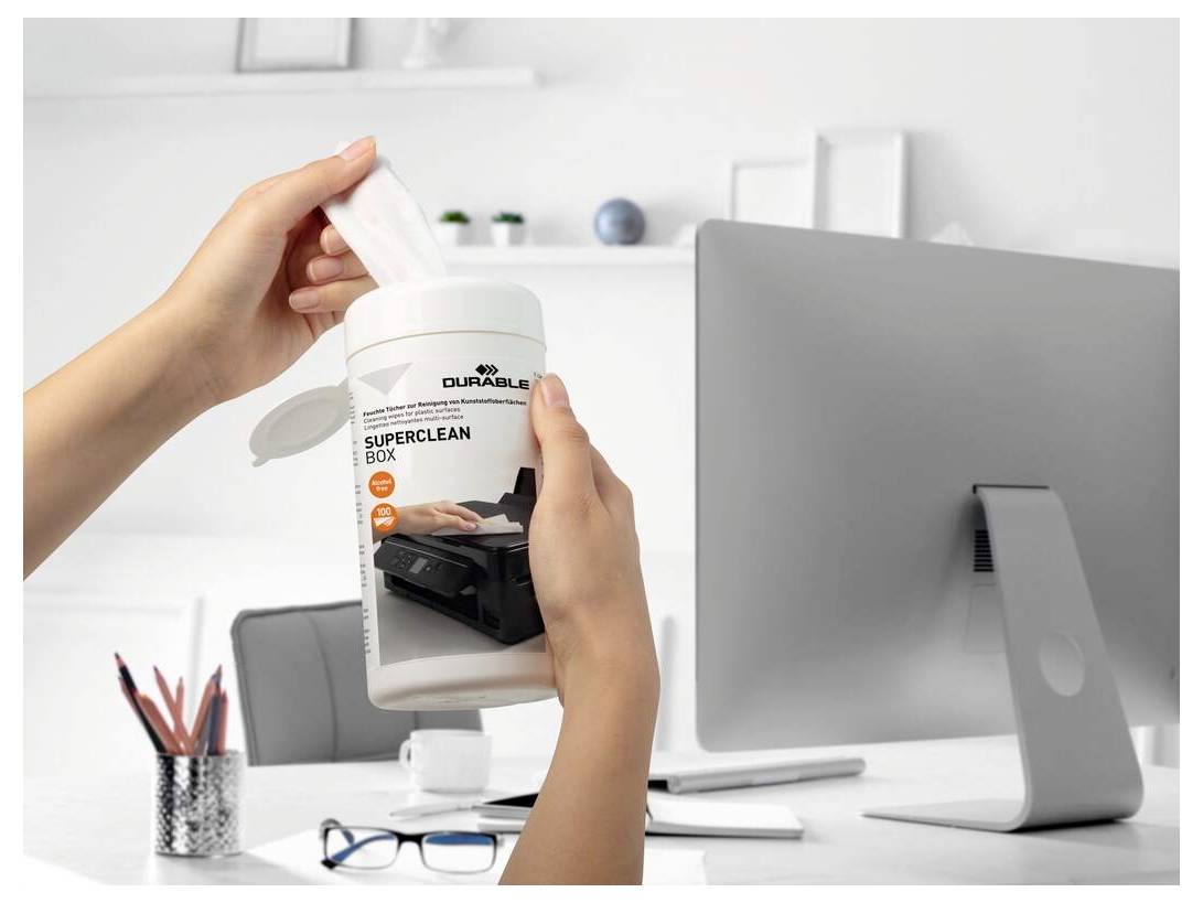 Les mains tenant un récipient étiqueté 'Boîte Superclean' dans un bureau avec un moniteur, des lunettes, un porte-stylos et des classeurs en arrière-plan.