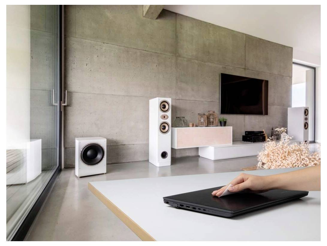 Un salon moderne au design minimaliste, avec un téléviseur, des enceintes et la main d'une personne reposant sur un ordinateur portable fermé sur une table.