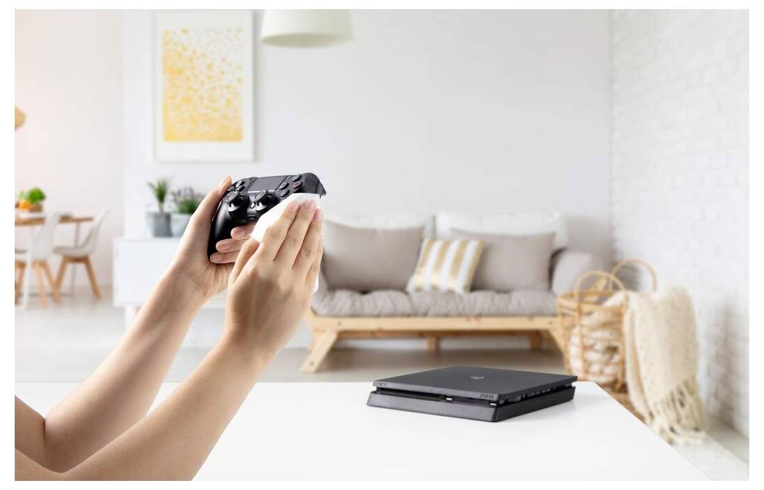 Une personne désinfecte une manette de jeu vidéo avec un tissu dans un salon lumineux, avec une console sur la table et un canapé en arrière-plan.
