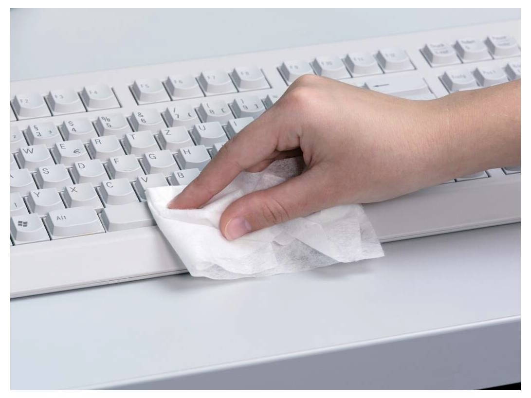 Une main nettoie un clavier d'ordinateur avec une lingette désinfectante blanche, mettant l'accent sur l'hygiène et l'entretien dans un environnement de bureau.