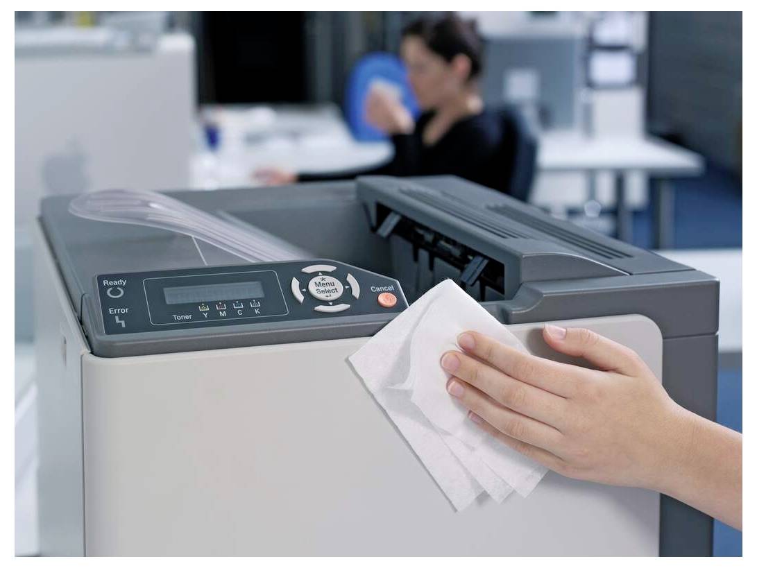 Une personne nettoie une imprimante de bureau avec un chiffon ; un employé de bureau flou est visible en arrière-plan.