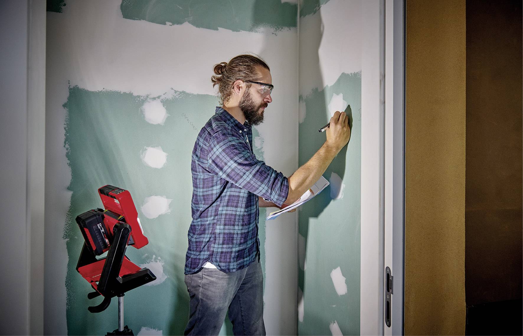 Un homme portant des lunettes écrit sur un presse-papiers, debout devant un mur enduit. Une lampe rouge est posée à côté. Scène de rénovation.
