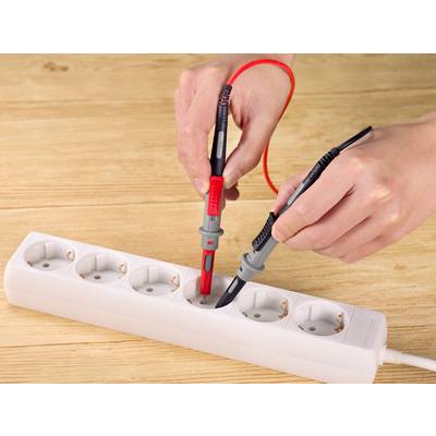 Une personne vérifie une bande multiprise blanche avec un testeur de tension sur une table en bois.