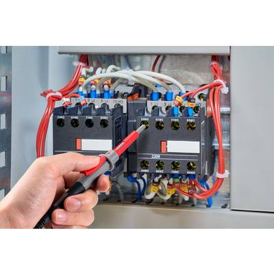 Une main tenant un tournevis vérifie les circuits électriques dans un armoire électrique ouverte. Des câbles de couleurs codées sont visibles.