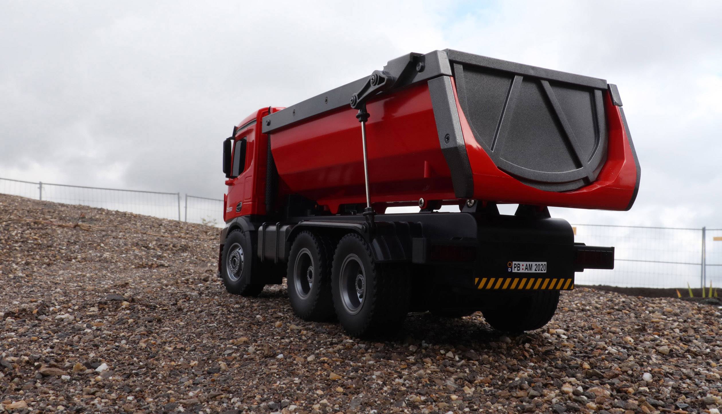 Un camion benne rouge est stationné sur une piste de gravier sous un ciel nuageux. Un grillage est visible en arrière-plan.