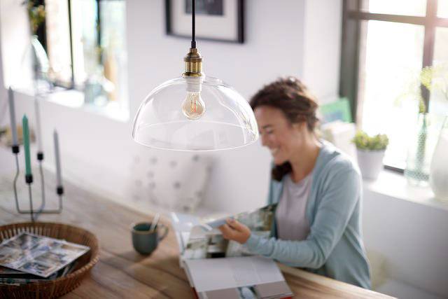 Une personne est assise à une table, souriant et feuilletant un magazine. Une ampoule avec un abat-jour en verre est suspendue au-dessus de la table.